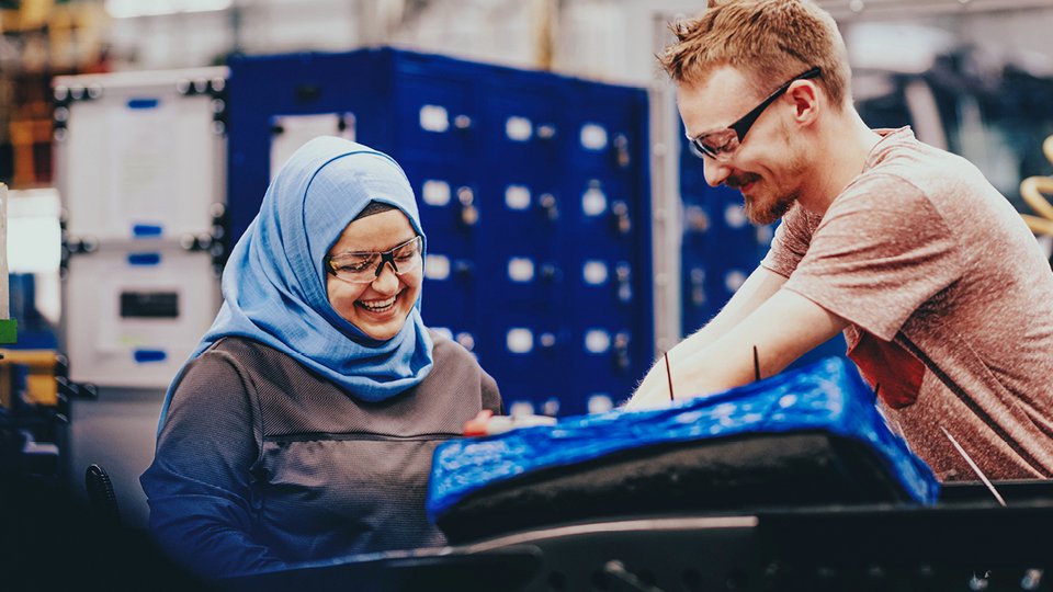 A woman with a headscarf and a man with safety goggles are smiling as they work together on a component in an assembly hall.