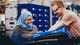 A woman with a headscarf and a man with safety goggles are smiling as they work together on a component in an assembly hall.