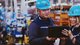 Two production employees wearing blue hard hats look at a process monitoring tablet in a busy production hall.