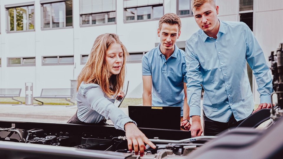 Three young students are working together on a vehicle frame in the open air. One student points to a technical component and holds a tablet while the other two students watch attentively.